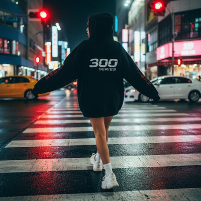 Person in oversized black 300 Series hoodie and sneakers crossing city street at night