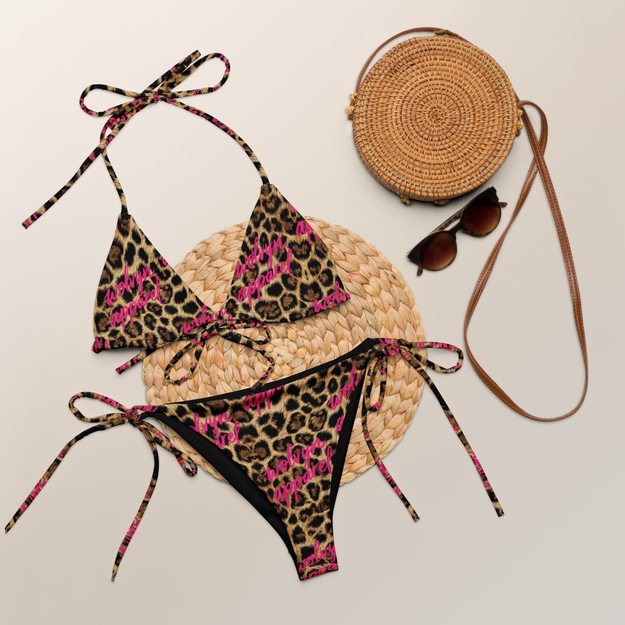 Leopard print string bikini set with pink lettering, straw bag, and sunglasses on neutral background.