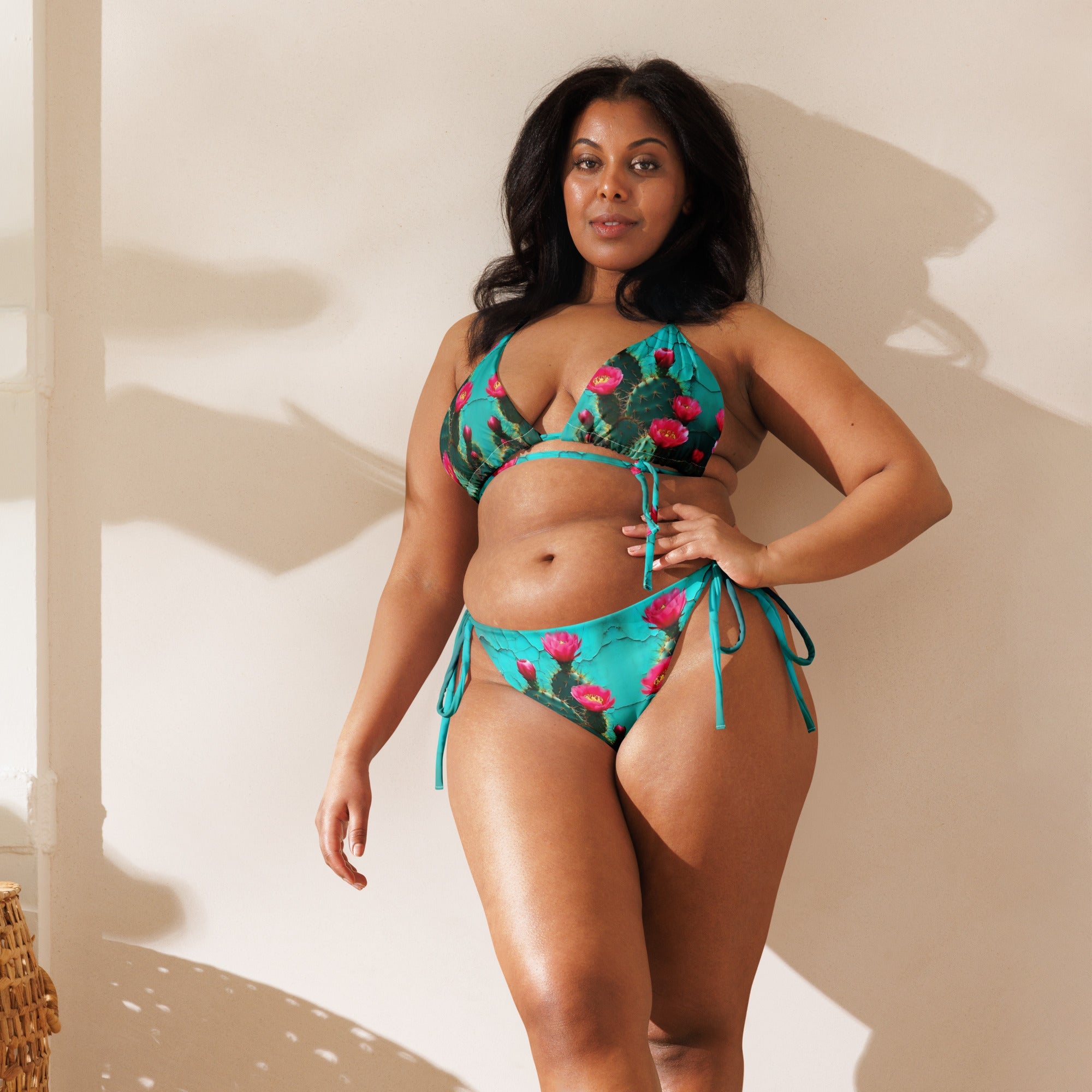 Woman wearing turquoise string bikini with red floral print, posing against a light wall
