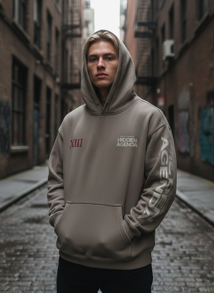 Man in an oversized taupe fleeced hoodie with Hidden Agenda text, standing in urban alley