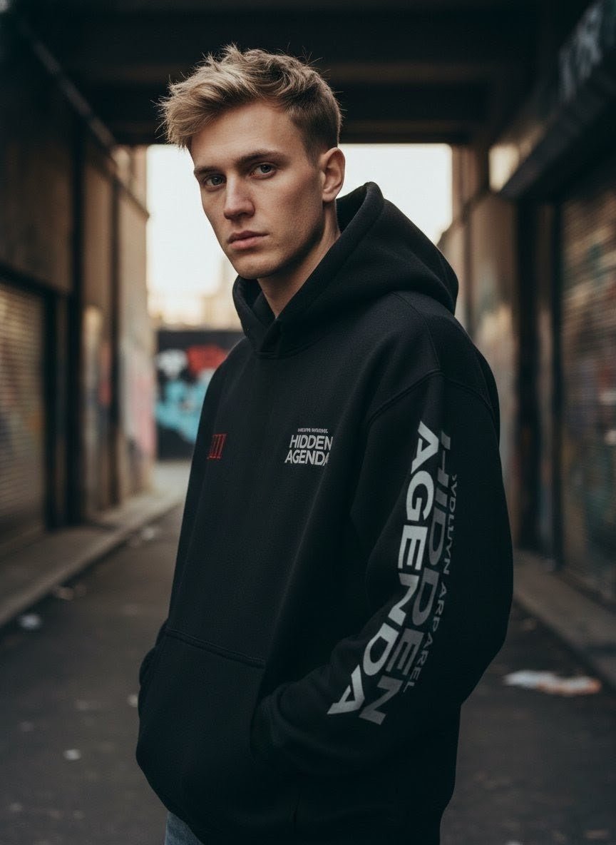 Man wearing black oversized fleeced hoodie with 'Hidden Agenda' text in urban alley