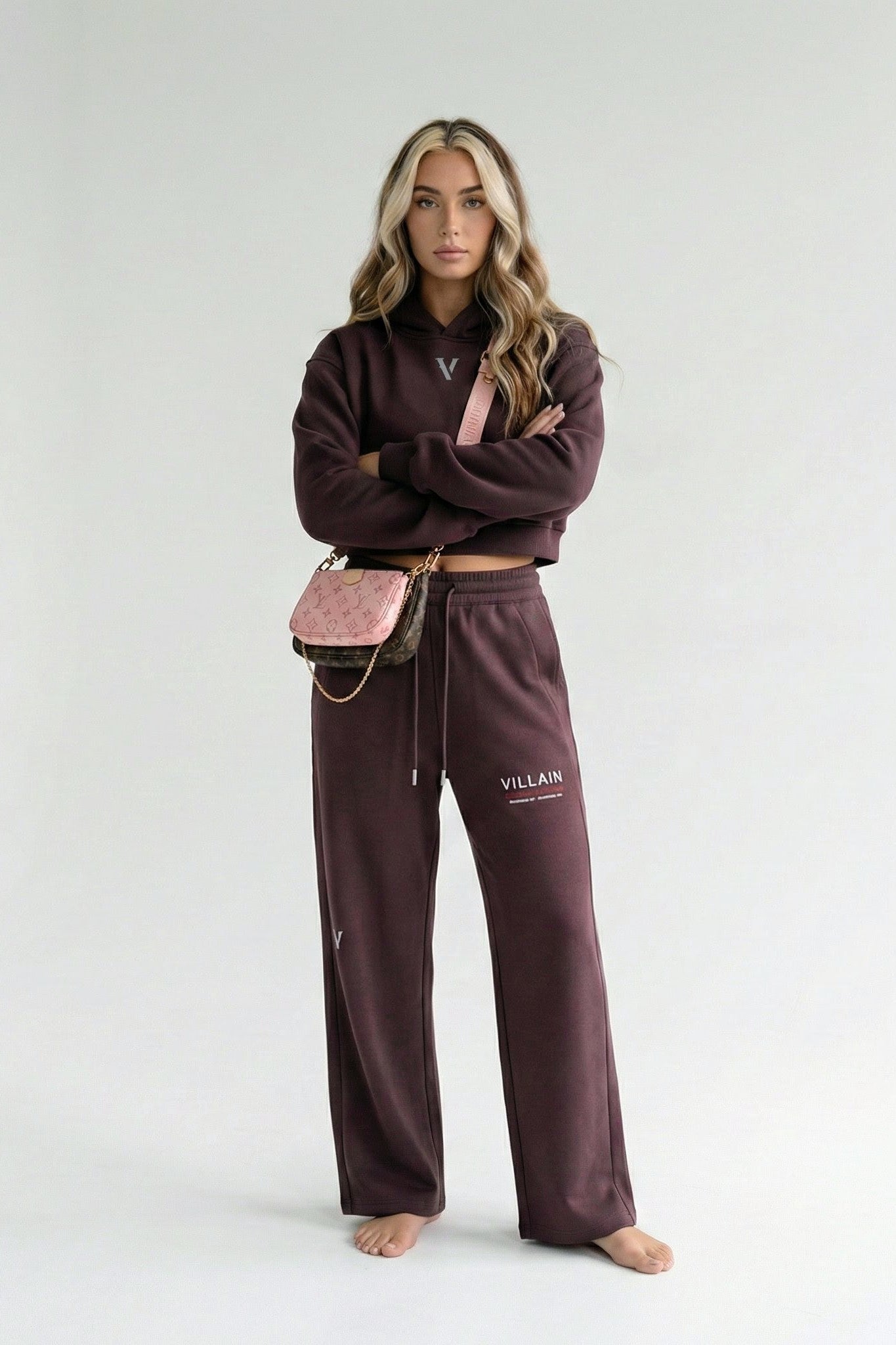 Woman in brown cropped hoodie and joggers lounge set with 'VILLAIN' print, barefoot, holding pink crossbody bag.