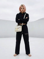 Woman in cropped black hoodie and sweatpants with 'Villain' text, barefoot, posing in a minimal white setting, carrying a white designer bag.