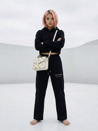 Woman in cropped black hoodie and sweatpants with 'Villain' text, barefoot, posing in a minimal white setting, carrying a white designer bag.