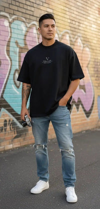 Man in black heavyweight knitted round neck T-shirt, ripped jeans, and white sneakers standing by a graffiti wall.