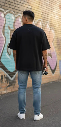 Man in black heavyweight round neck t-shirt, blue jeans, and white sneakers, standing outdoors by a graffiti wall.