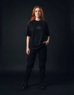 Woman wearing a black heavyweight knitted round neck t-shirt with 'Villain' text, black pants, and boots, posing against a dark background.