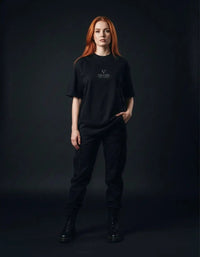 Woman wearing a black heavyweight knitted round neck t-shirt with 'Villain' text, black pants, and boots, posing against a dark background.