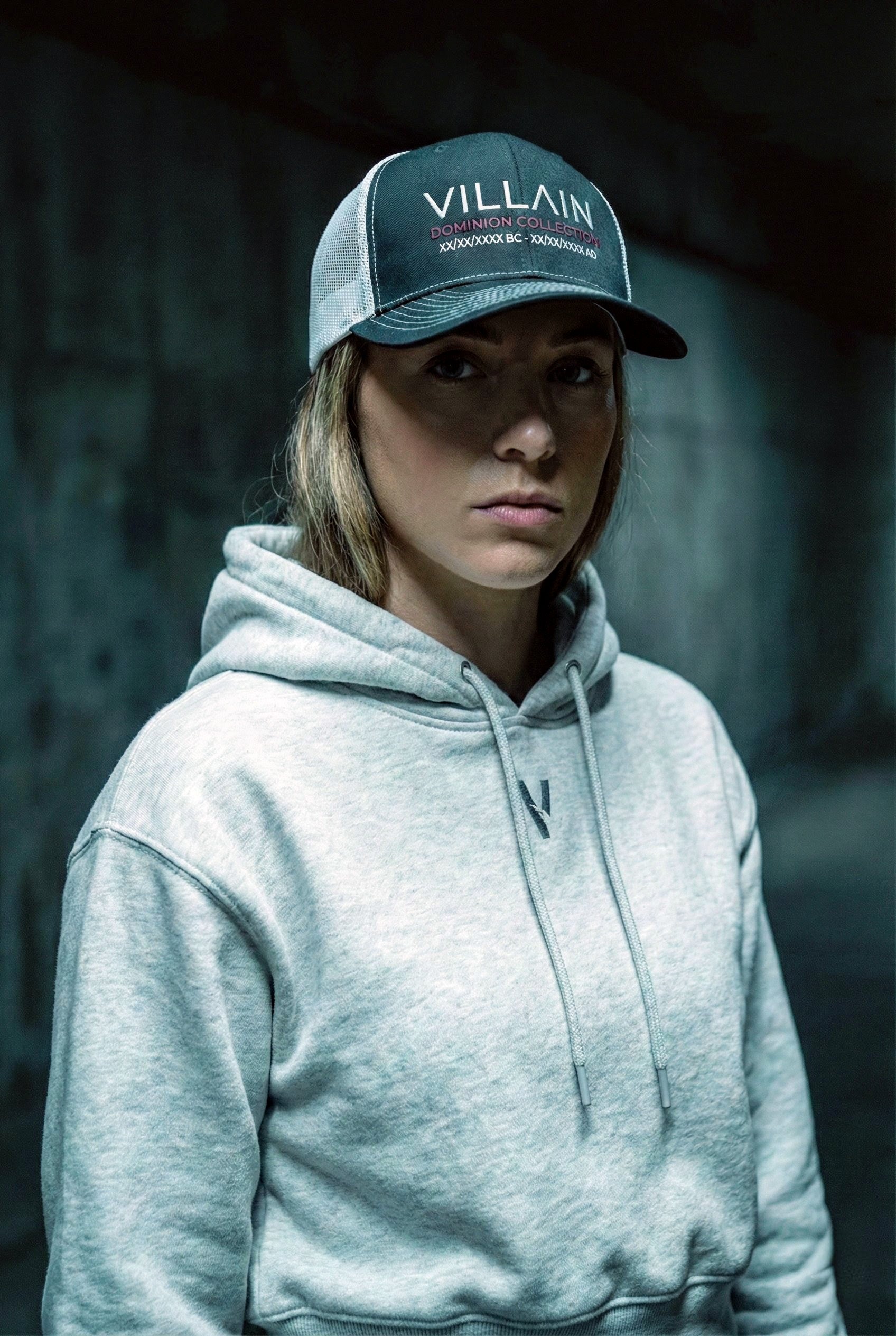 Woman wearing a Villain Dominion Collection trucker cap and light gray hoodie in a moody urban setting