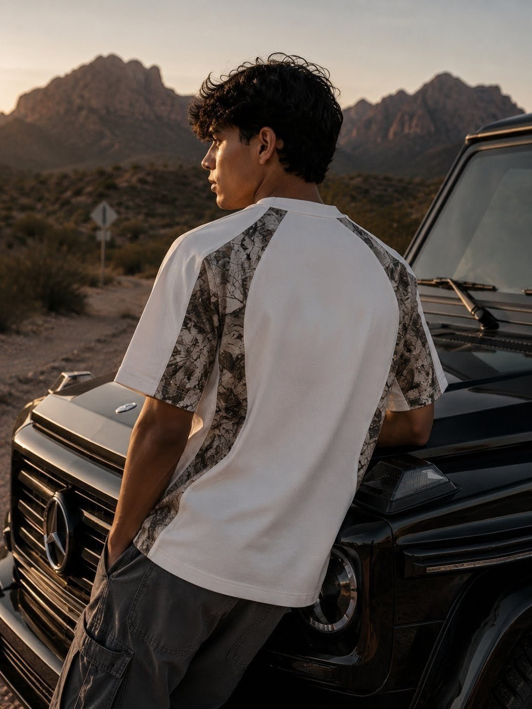 Man in casual streetwear t-shirt with camo panels leaning on black SUV in desert landscape