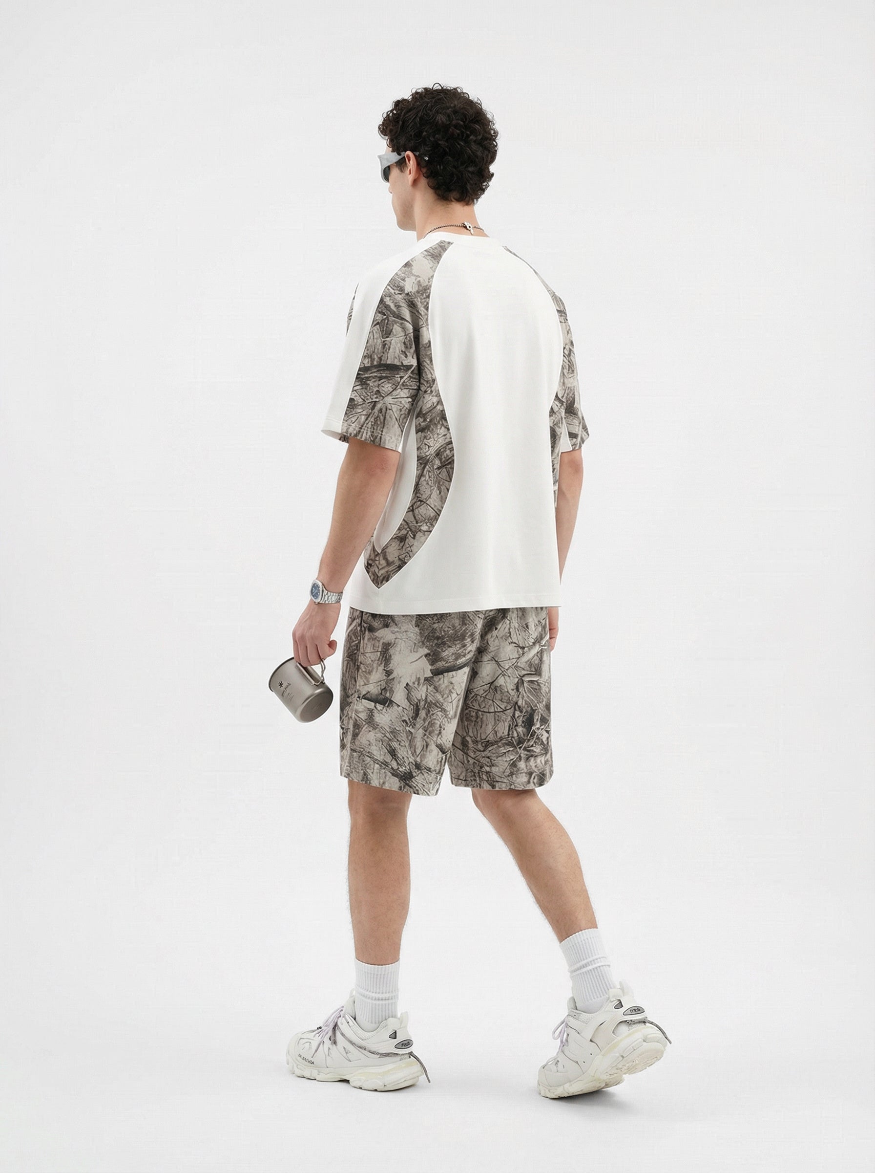 Man wearing camouflage panelled t-shirt and shorts, holding a mug, in a studio