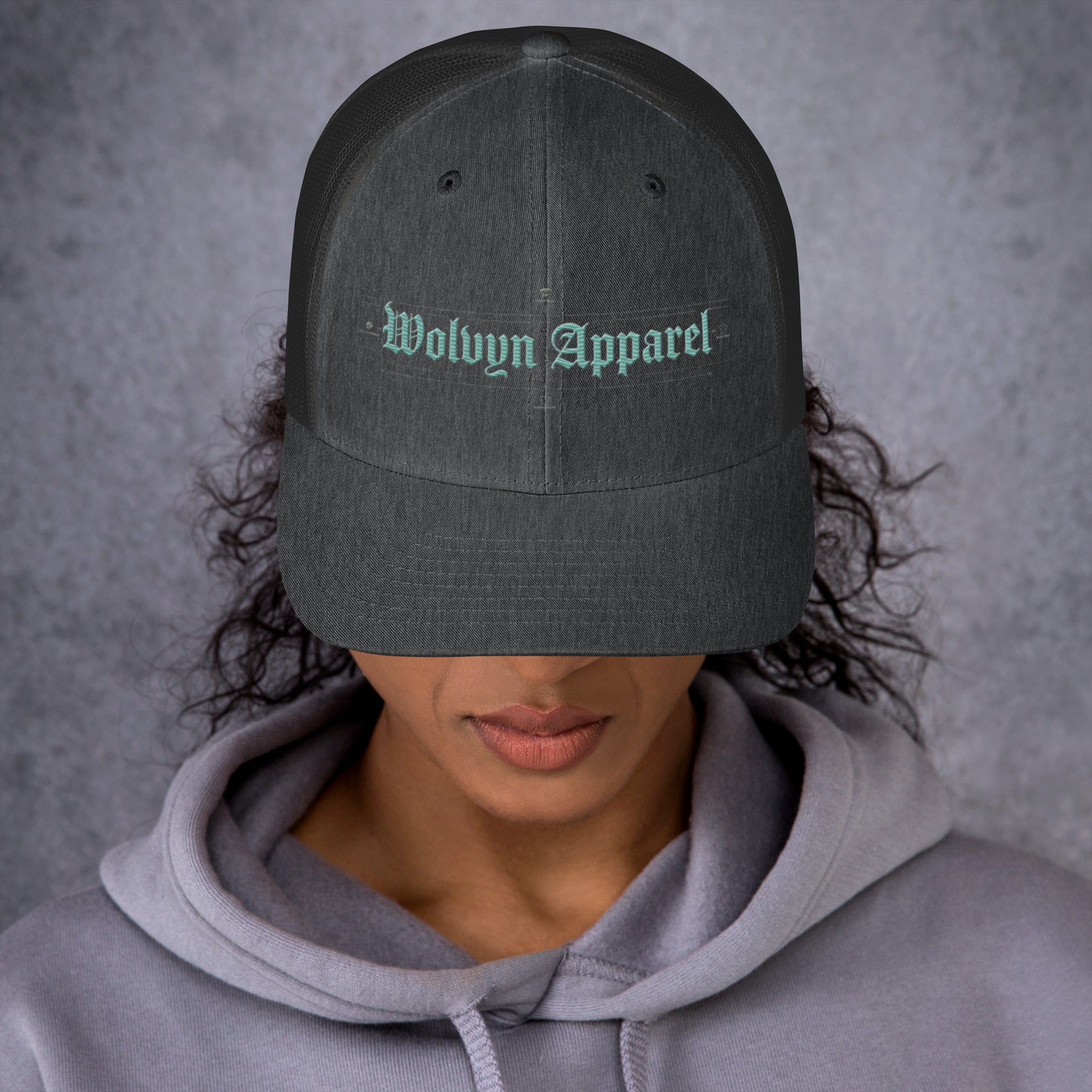 Mid-profile charcoal embroidered trucker cap with mesh back, worn by woman in gray hoodie.
