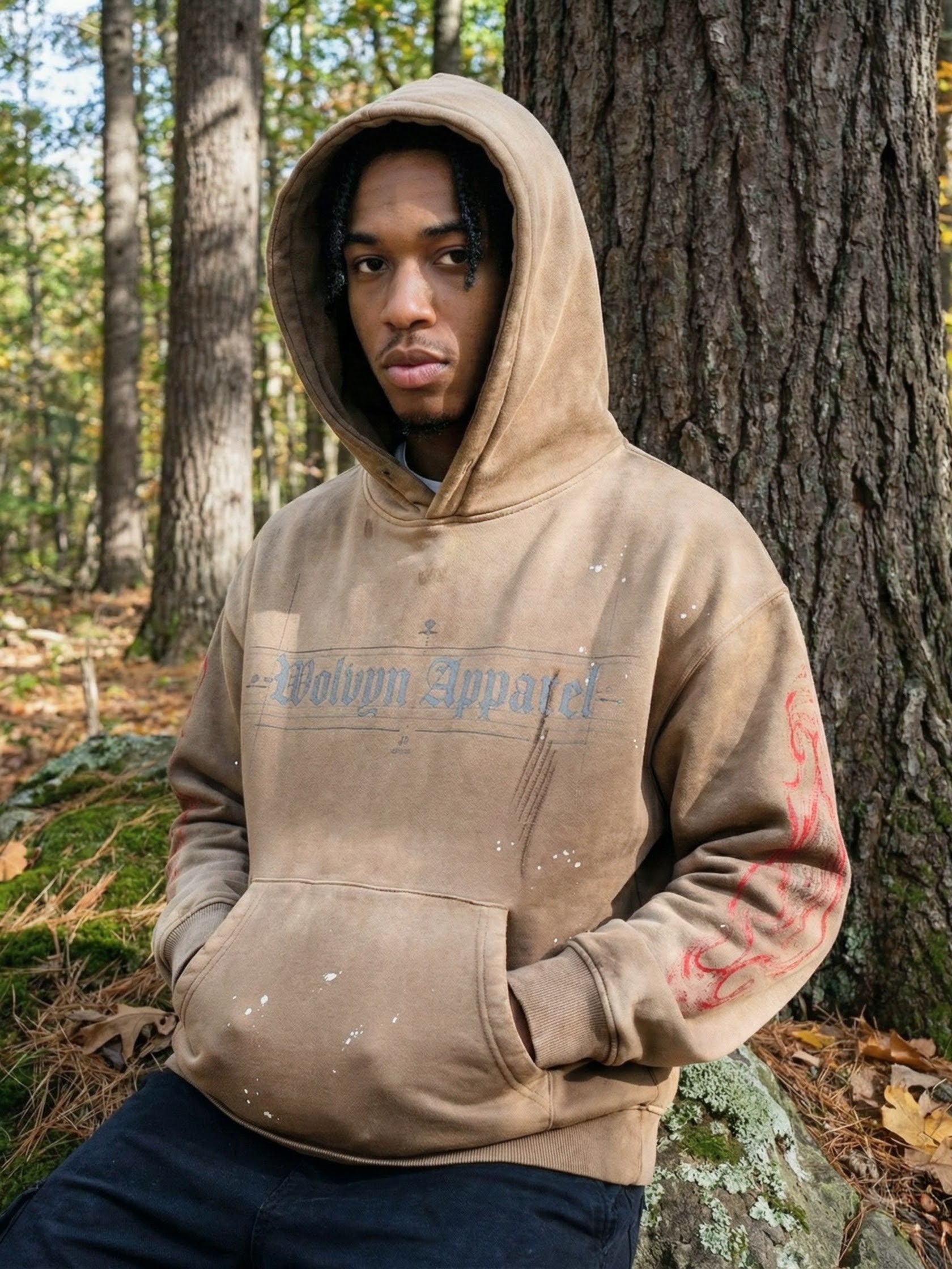 Man wearing a tan weathered fleece hoodie with frayed details and red sleeve print in the forest