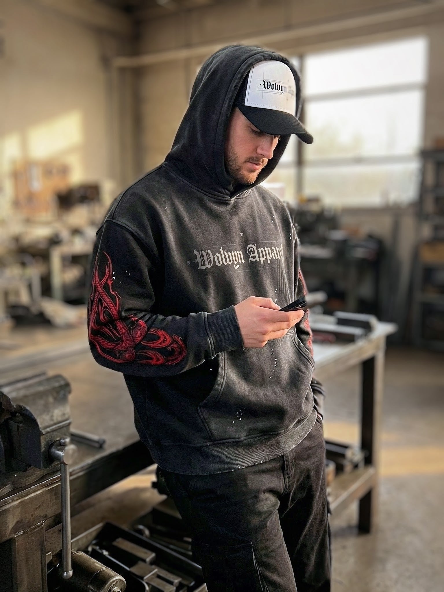 Man wearing distressed black fleece hoodie with graphic sleeves and logo cap in industrial setting