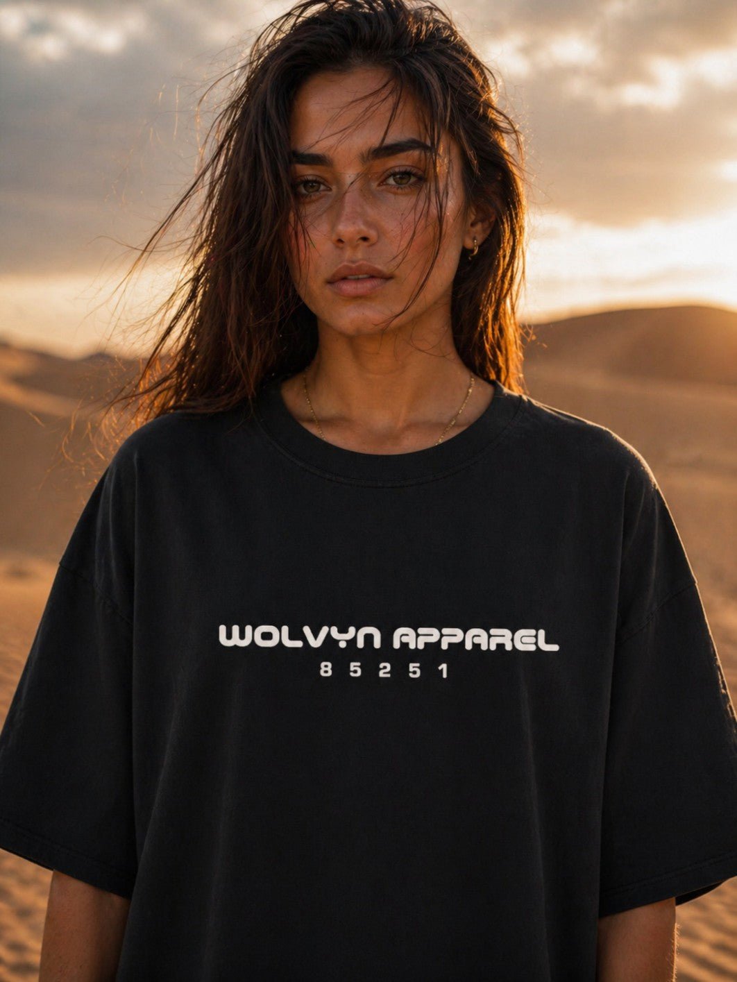Woman in a black heavyweight boxy T-shirt with 'Wolvyn Apparel 85251' text, standing in desert at sunset.