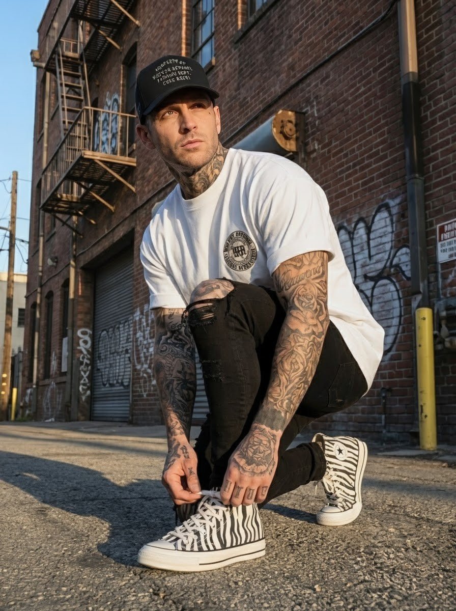 Tattooed man in streetwear tying zebra sneakers, wearing white logo tee and black five-panel cap outdoors