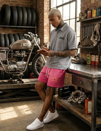 Man in a grey heavyweight snow-washed polo t-shirt and pink shorts, standing in a garage workshop next to a Triumph motorcycle, taking notes.