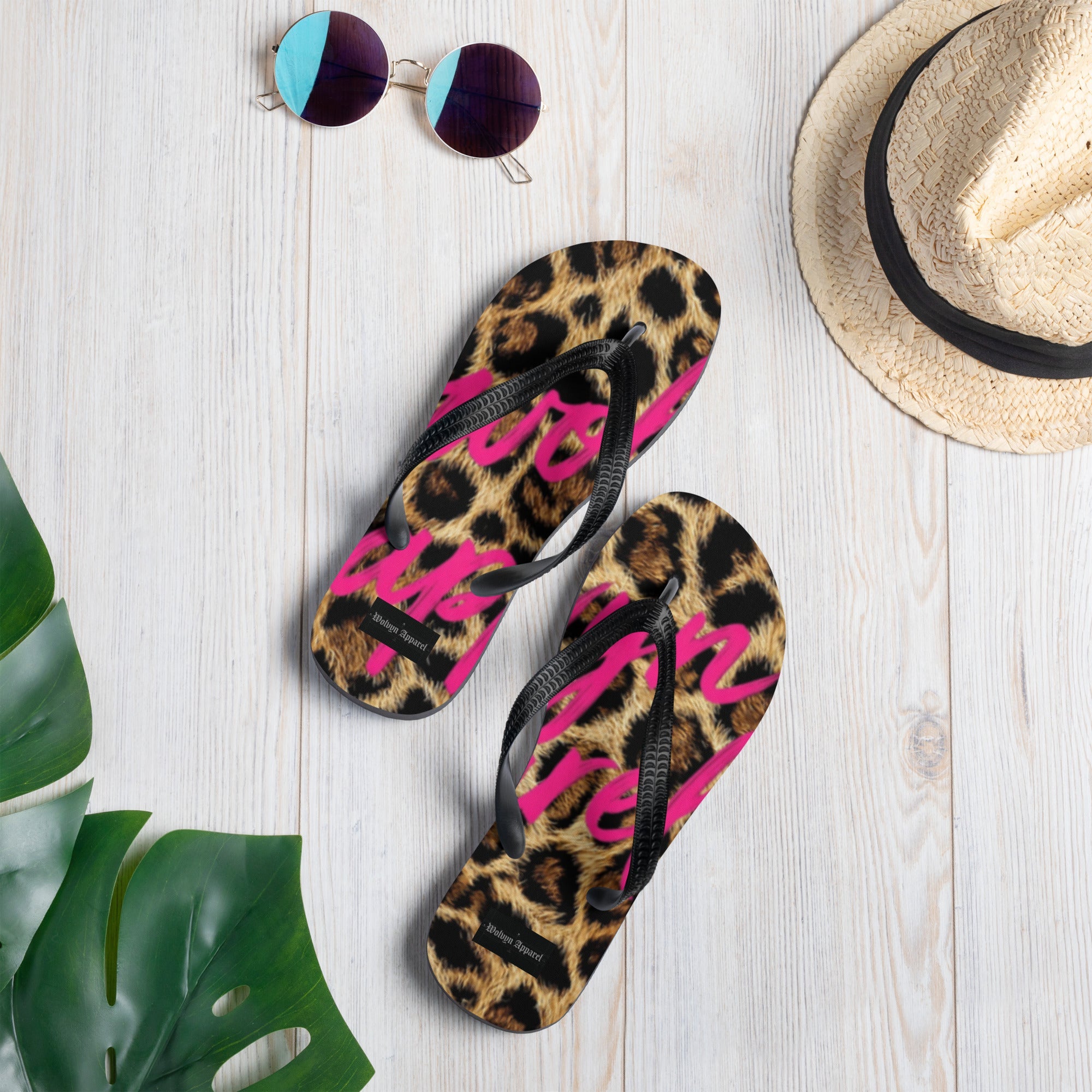 Leopard print flip flops with bold pink script on a light wood surface, styled with sunglasses, straw hat, and tropical leaves.