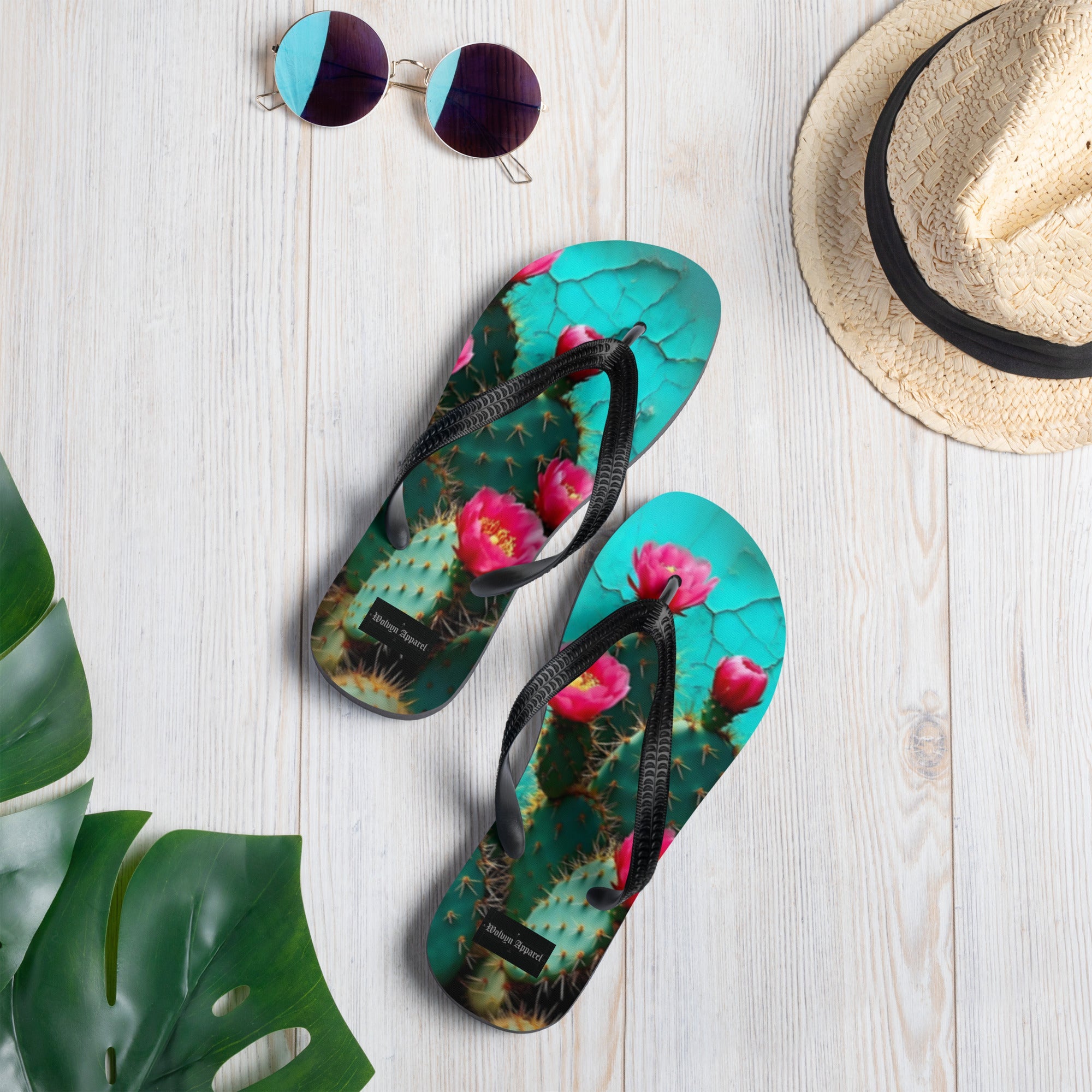 Flip flops with cactus and pink flower design on light wood floor, summer hat and sunglasses nearby