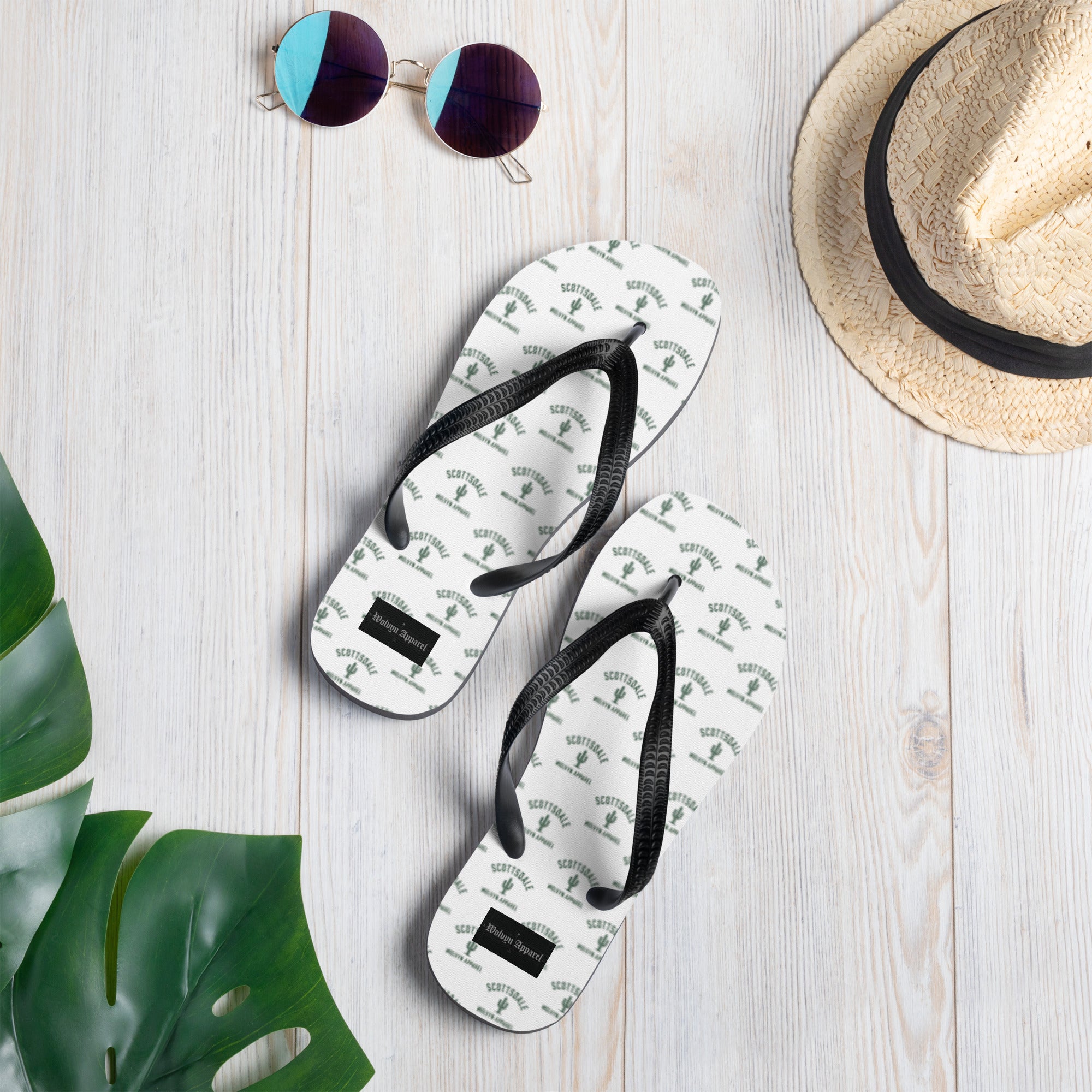 White flip flops with black straps, cactus and 'Scottsdale' print, on a wooden floor with hat and sunglasses.