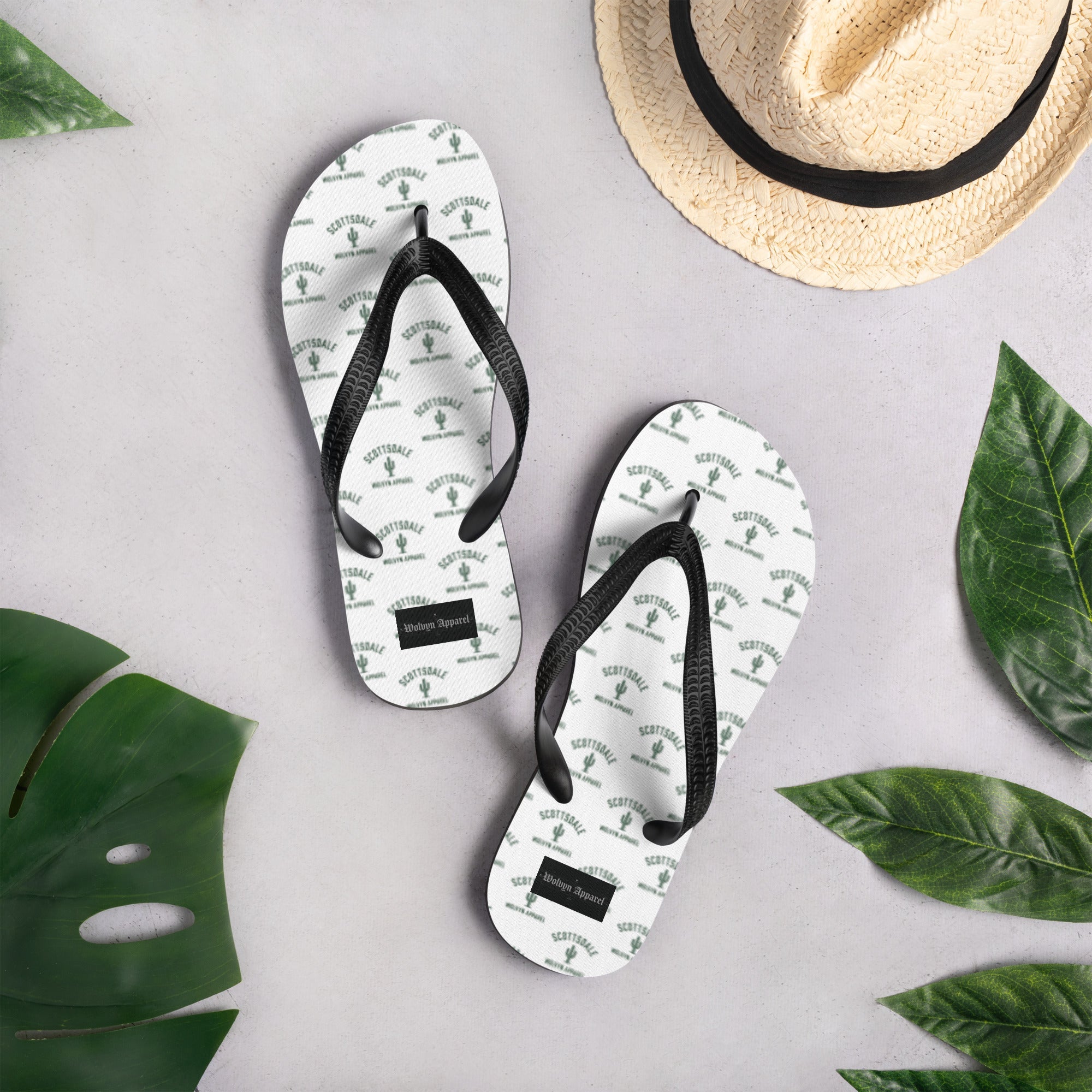 White flip flops with Scottsdale cactus print, black straps, styled with tropical leaves and a straw hat