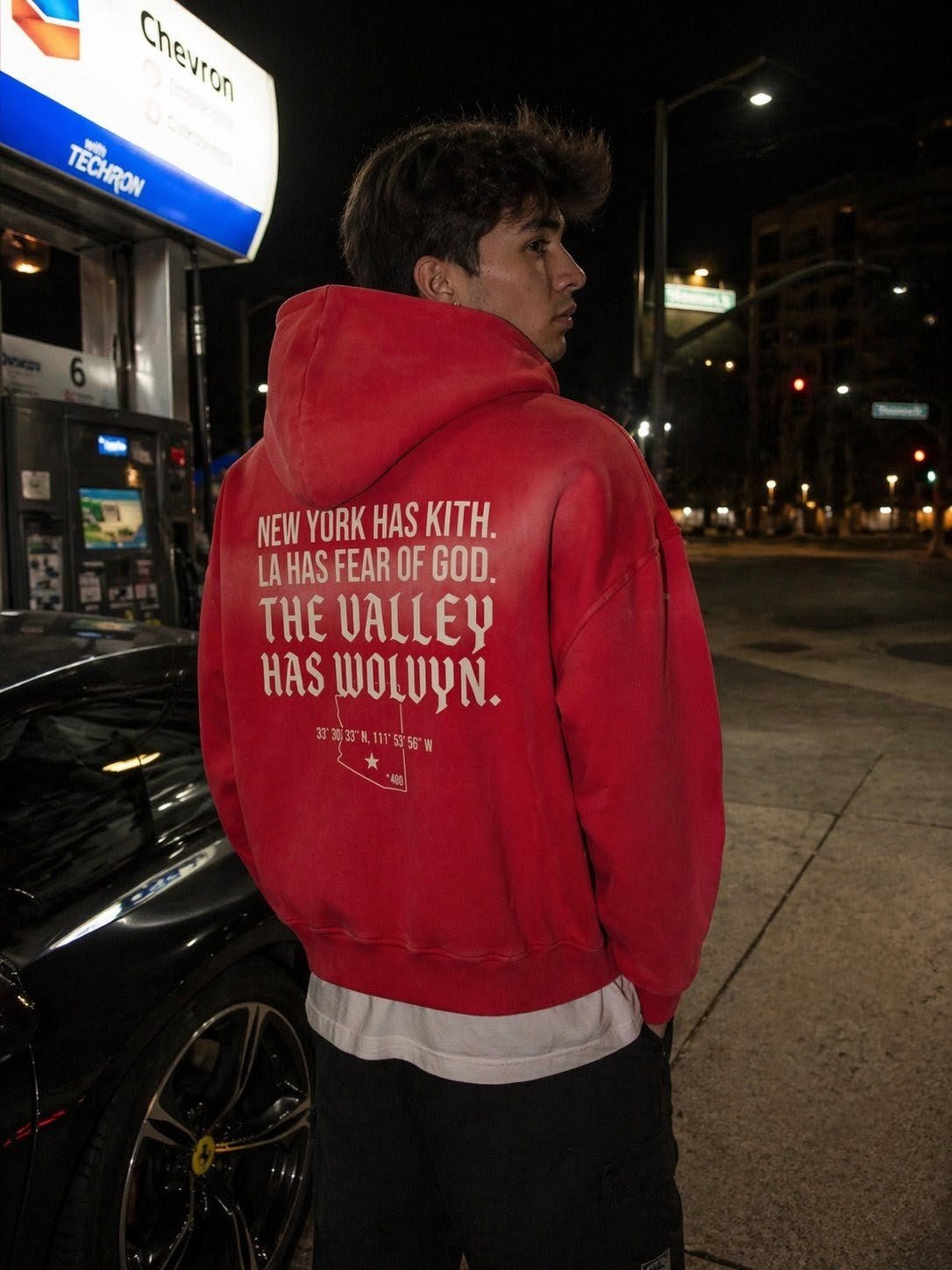 Man wearing red fleece hoodie with white graphic text at night near a gas station on street.