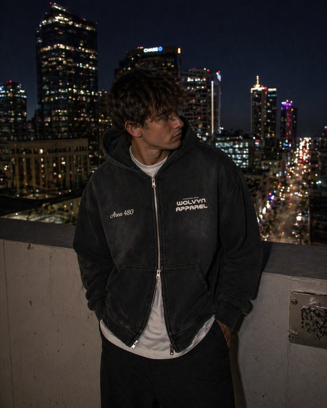 Man wearing faded black boxy fleece hoodie with two-way zipper and text details, city skyline at night