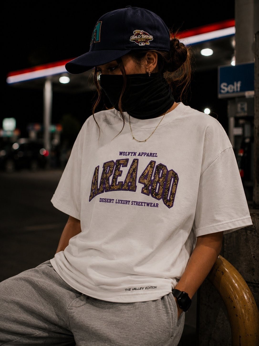 Person in casual streetwear at night, wearing AREA 480 white t-shirt and cap near gas station