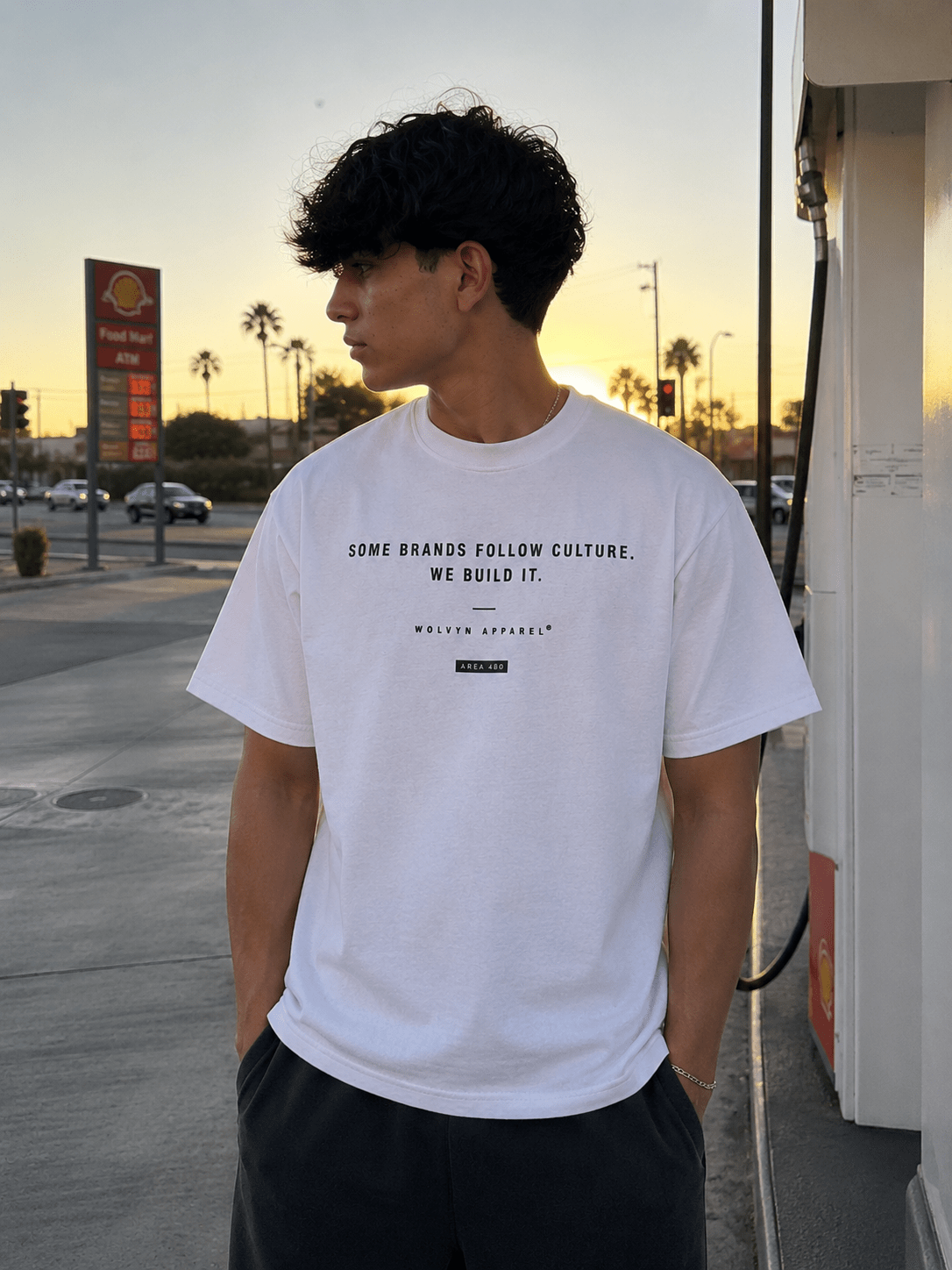 Young man wearing a white graphic streetwear t-shirt at a gas station during sunset.