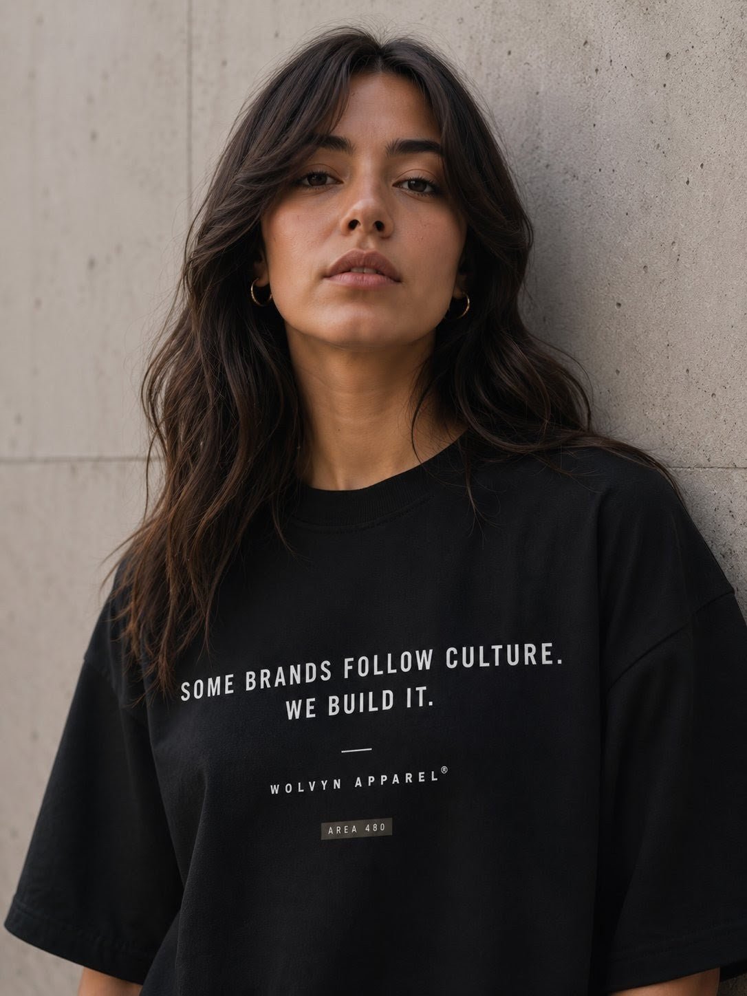 Woman in black graphic streetwear t-shirt with bold white text, posing against concrete wall