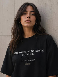 Woman in black graphic streetwear t-shirt with bold white text, posing against concrete wall
