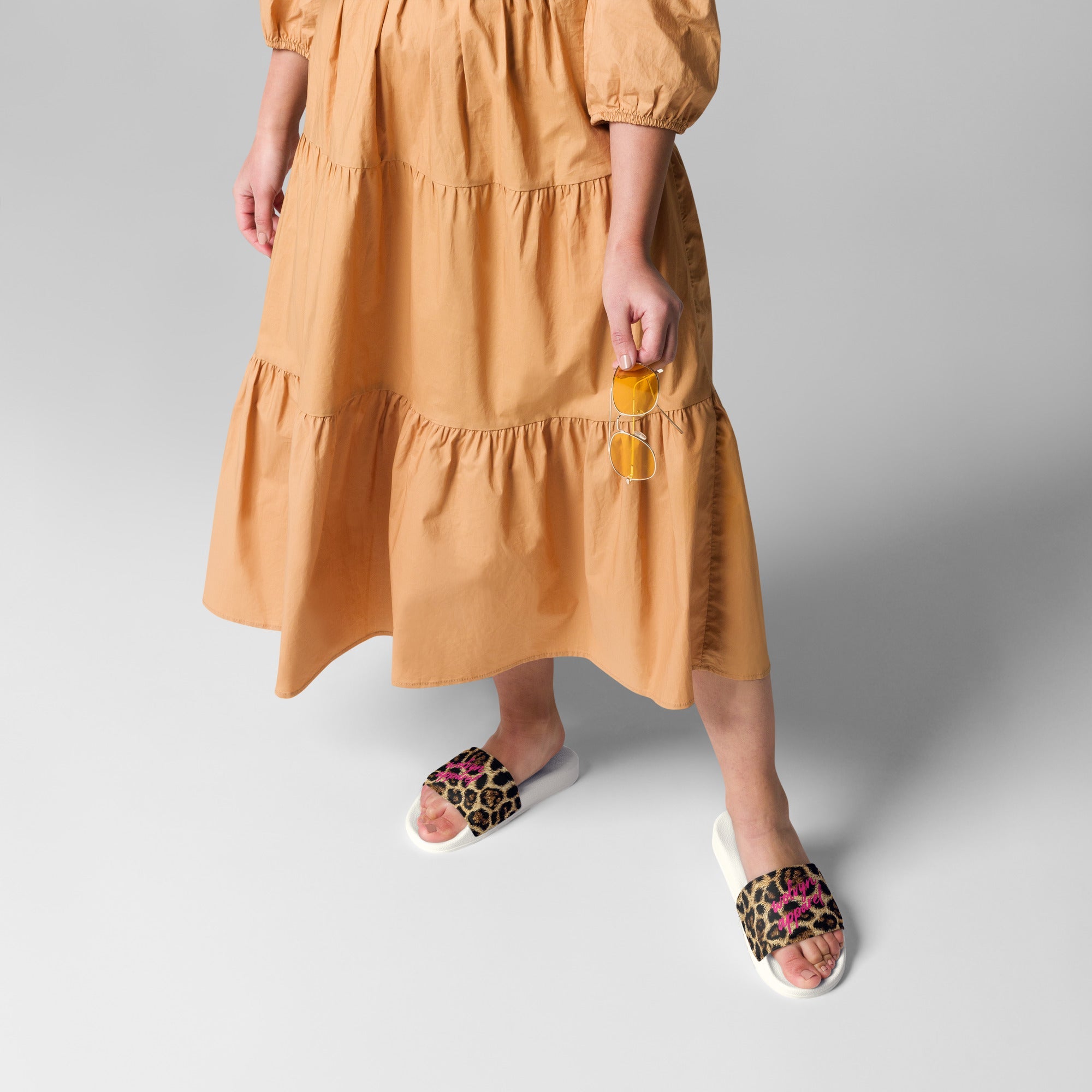 Woman in tan dress wearing leopard print slides and holding yellow sunglasses