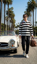 Man in a black and white color block stripe polo shirt and joggers, walking beside a vintage convertible on a palm-lined street.