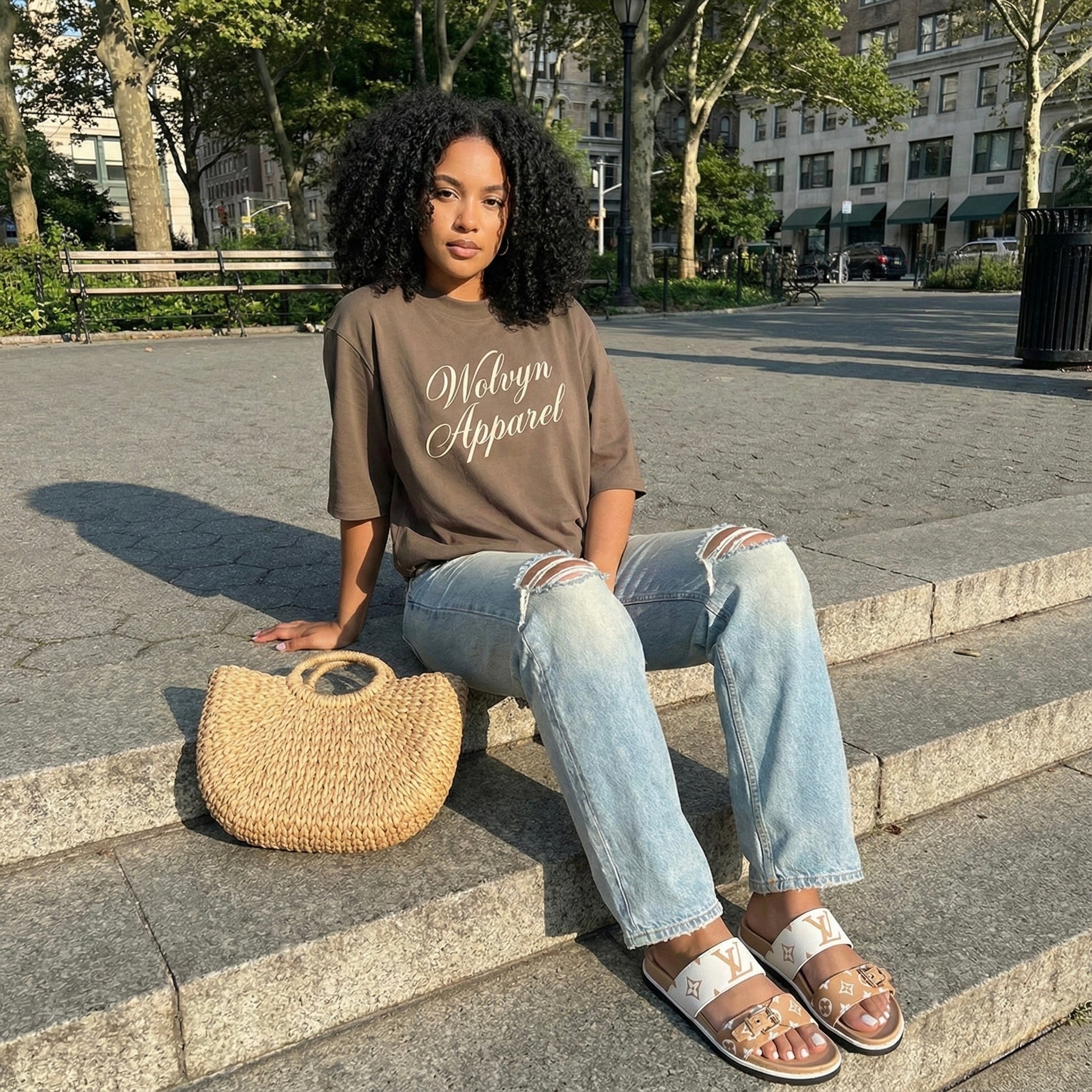Woman sitting outdoors wearing a brown graphic t-shirt, ripped jeans, and sandals, with a woven bag.