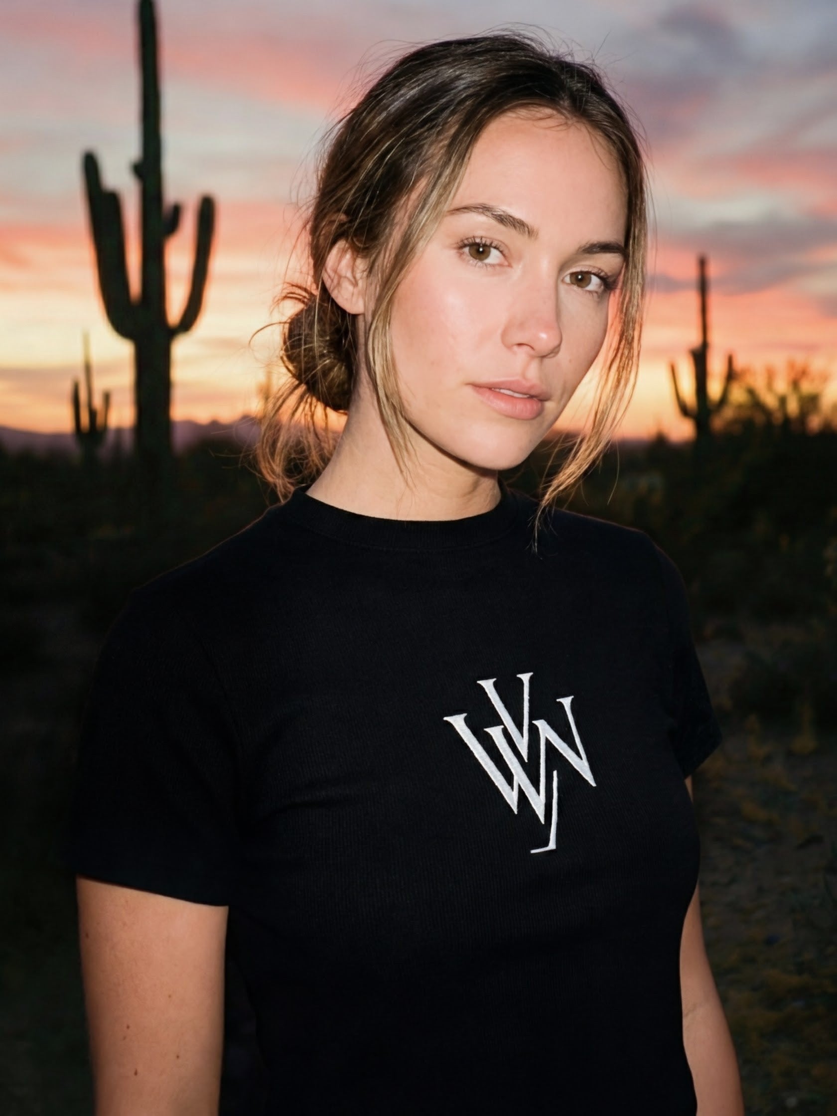 Woman wearing black slim fit mock neck t-shirt with white monogram, desert sunset background