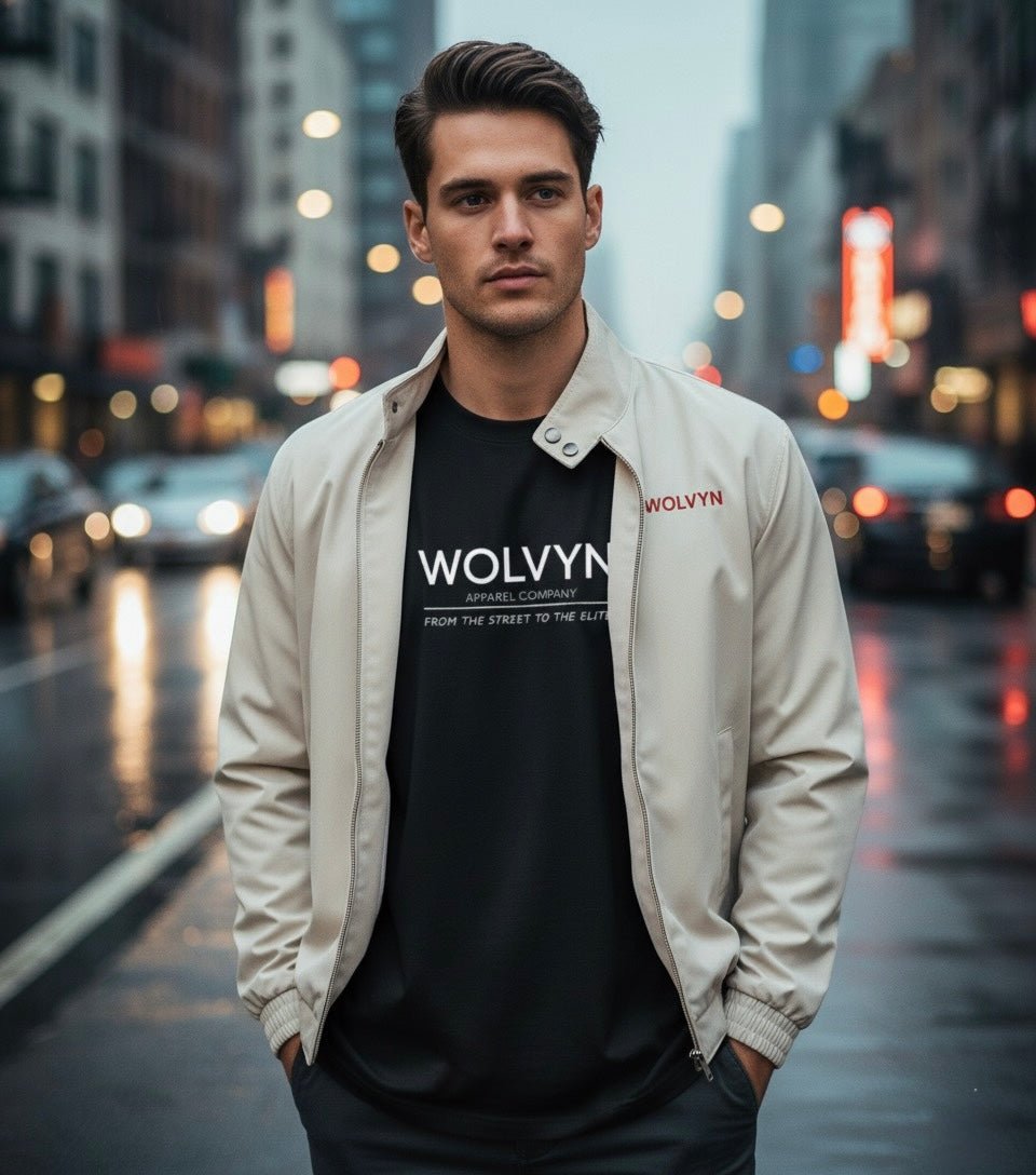 Man wearing cream stand collar bomber jacket over graphic tee on a city street at dusk.